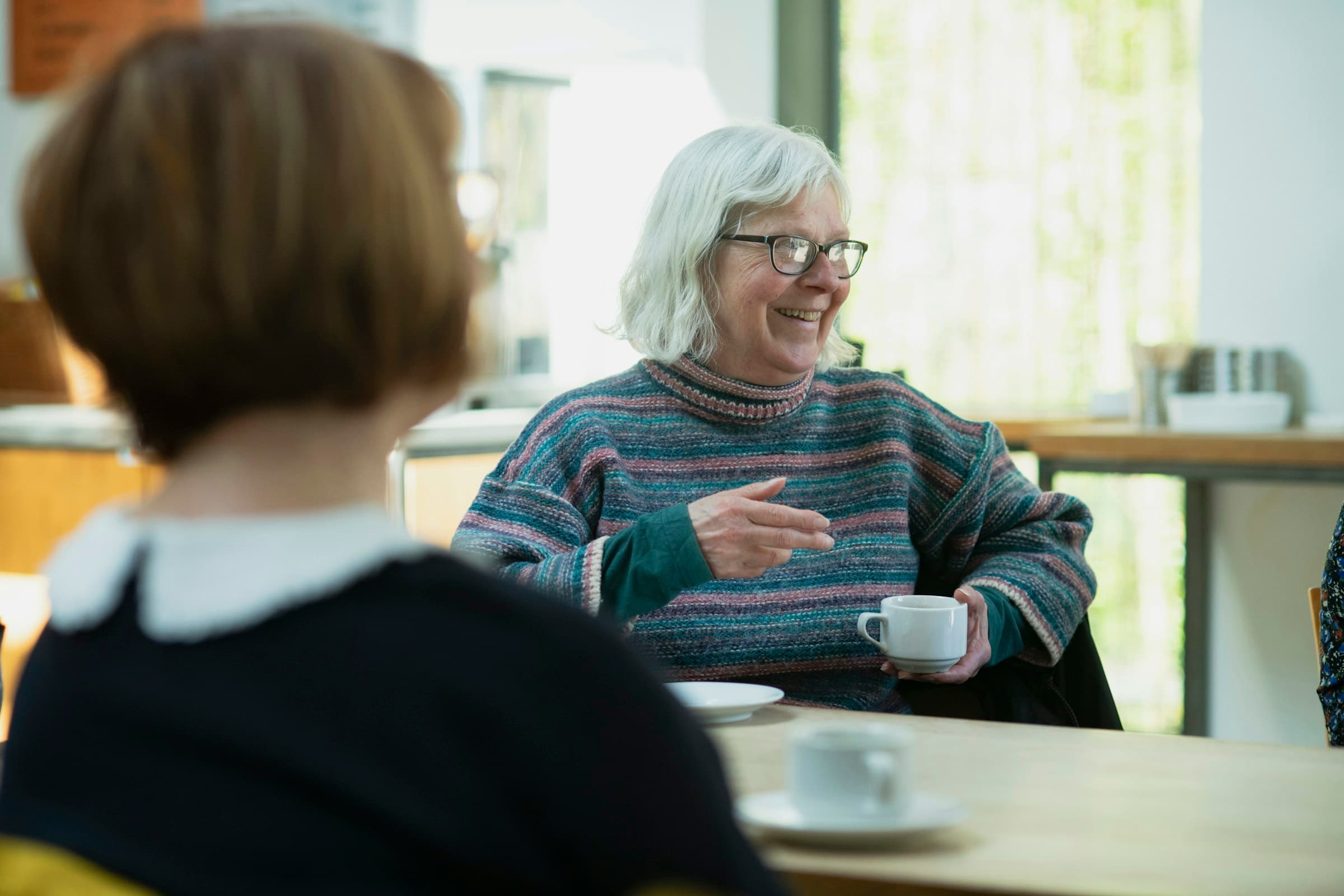An older woman laughing over a cup of tea, sitting across a kitchen table from a younger woman.