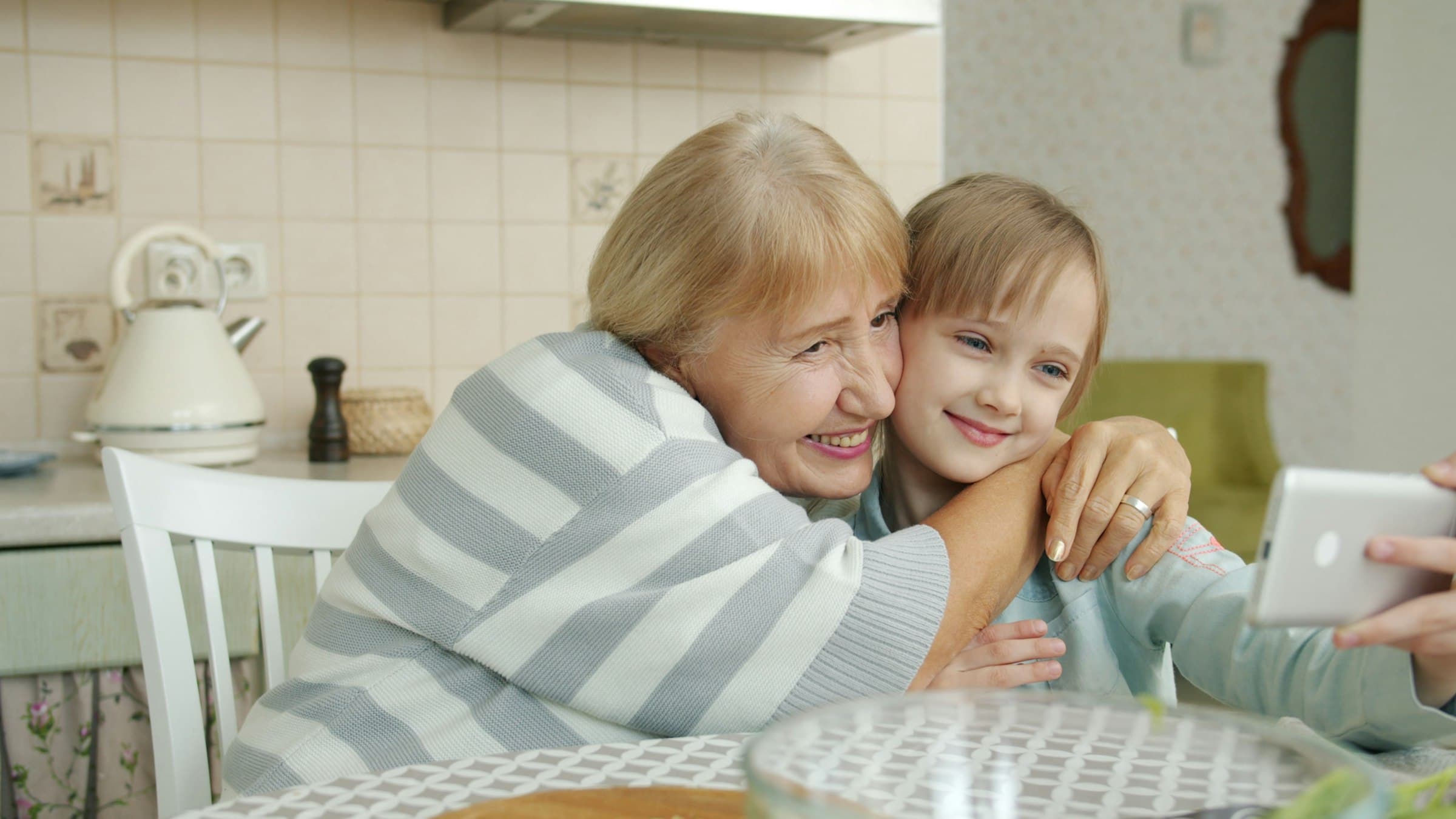 A grandmother and her granddaughter at a kitchen table, cheek to cheek and laughing, holding up a phone.
