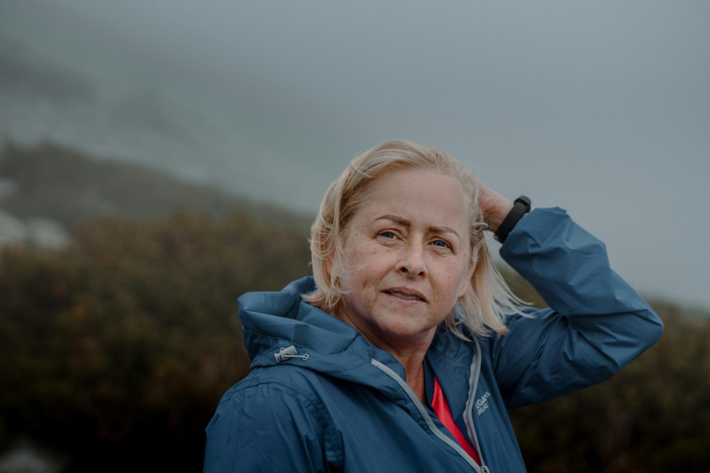 A woman in her sixties in a blue jacket, standing outdoors on open hillside on an overcast day, relaxed and smiling.